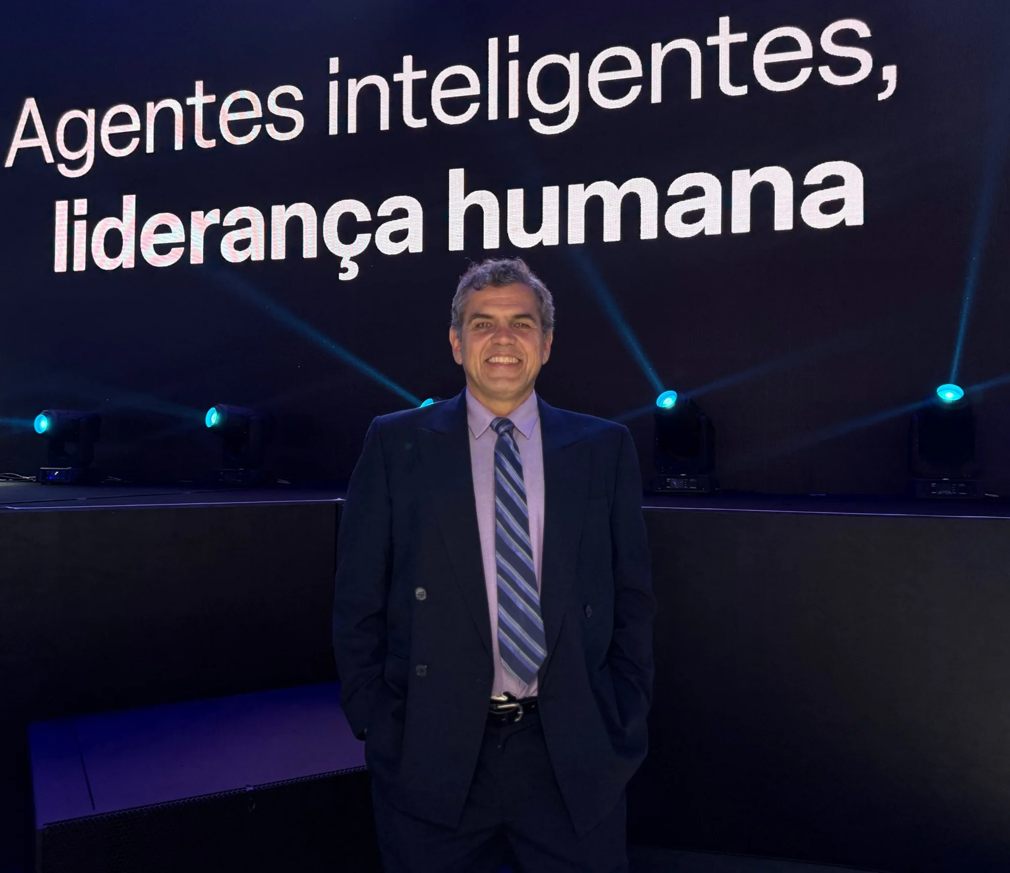 Dorian Lacerda in a suit, smiling, standing on a stage; behind him an LED wall reads “Agentes inteligentes, liderança humana” — intelligent agents, human leadership